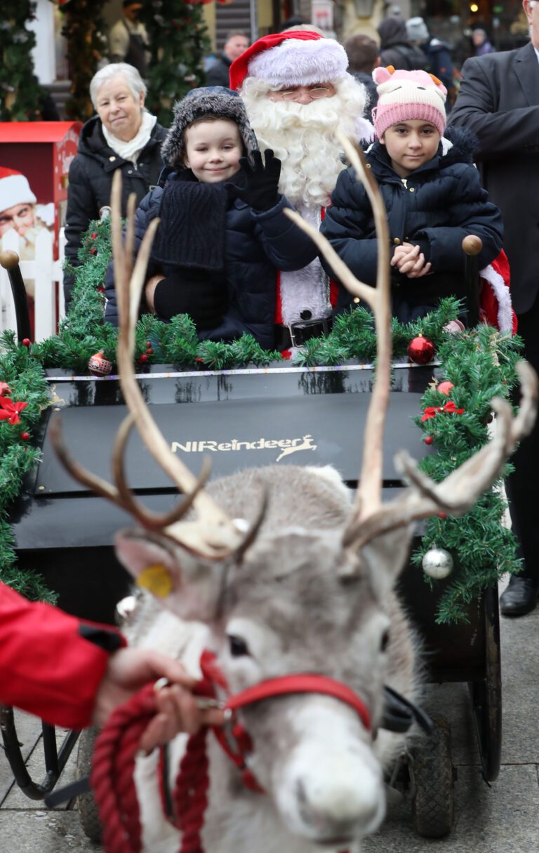 Children enjoying a charity ride sit with Santa in a festive sleigh, pulled by a majestic reindeer adorned with antlers, while smiling onlookers cheer in the background.