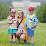 A woman crouches on the grass, smiling with two children. They are wearing sporty clothes and numbered race bibs, set against the scenic backdrop of trees and the iconic Stormont building in the distance.