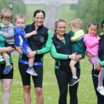 Six women in athletic attire each holding a child, standing outdoors on a grassy path lined with trees, like they're gearing up for the Stormont run.