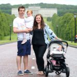 A family of four stands on a wide road leading to Stormont. One parent holds a young child, while the other stands by a stroller with a baby. A large building and trees provide a picturesque backdrop.