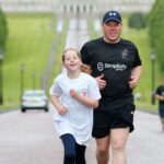 A man and a young girl are jogging on a paved path with the grand Stormont building in the background. Both are smiling.