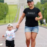 An adult and child are walking hand-in-hand on a paved path outdoors, with the majestic Stormont building in the background. The adult wears a black cap and shirt emblazoned with "Believe in Yourself," creating an inspiring moment amidst iconic scenery.