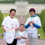Three people wearing "CCUC Stormont Mile" shirts stand proudly on a path, with the grand backdrop of the Stormont building. One of them holds a shoe-shaped sign, capturing the spirit of their run.