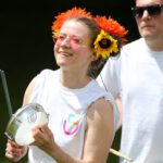 At an outdoor event near Stormont, a person wearing sunglasses and floral headpieces plays a handheld drum. In the background, another individual, donning a white shirt, joins in the vibrant atmosphere.