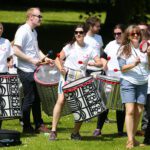A group of people in matching white shirts play percussion instruments outdoors on a sunny day, creating a lively atmosphere reminiscent of the vibrant gatherings often seen at Stormont.