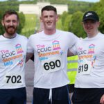Three men wearing race bibs and matching event shirts stand together outdoors, with the iconic backdrop of Stormont adding to the excitement of the day.