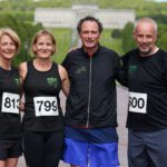 Four people in running gear with numbered bibs pose outdoors on a path lined with trees, the grand Stormont building visible in the background.