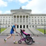 A man in running gear pushes a child in a wheelchair past Stormont's grand facade, blending athleticism with history.