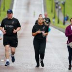 A group of four people jogging along the wide, paved road near Stormont, lined with lush grass. They are wearing casual athletic clothing.