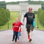 A man and two children run along a wide path lined with trees, Stormont's grand facade visible in the background. They wear casual clothes and appear to be enjoying the outdoors.