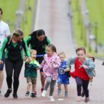 A group of adults and children in athletic attire are joyfully running along a pathway, set against the picturesque backdrop of Stormont.