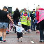 A woman and a toddler in matching shirts walk hand in hand at an outdoor event near Stormont. People are gathered around, some wearing high-visibility vests.