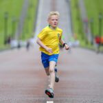 A boy in a yellow soccer jersey and blue shorts runs on a wide path through Stormont, with blurred figures and lush greenery in the background.