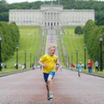 A boy in a yellow shirt runs along the path leading to Stormont, surrounded by lush green lawns and people enjoying the background scenery.