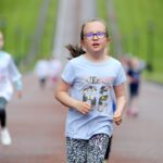 A young girl with glasses and a blue shirt runs energetically on a paved path near Stormont, with other children playing in the background.