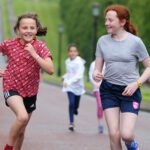 Two girls are running energetically along a paved path at Stormont, with others in the background. One wears a red shirt and the other a gray one, both radiating happiness as they enjoy their outdoor adventure.