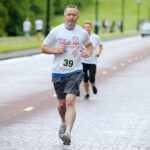 A man in a white T-shirt and shorts runs on the paved road during the Stormont race, with bib number 39, while other participants follow closely in the background.