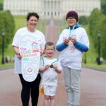 Three people in matching white T-shirts stand on a pathway at Stormont, proudly holding medals and a sign that reads "The CCUC Stormont Mile," with a large building in the background.