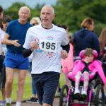 A man wearing race number 195 is running on the Stormont track. Nearby, a group of people, including a person in a wheelchair, are watching intently.