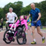 A man in blue pushes a young girl in a wheelchair through the Stormont race event. Another participant, wearing a race bib, walks in the background with a phone in hand, while spectators line the route.