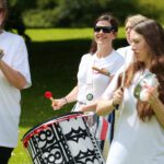 A lively group of people playing percussion instruments outdoors, all donned in white shirts, with the iconic Stormont building looming in the background. One woman enthusiastically beats a large drum while smiling broadly.