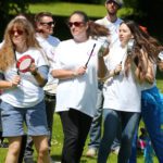 On a sunny day at Stormont, a group of people in white t-shirts gather outdoors, joyously playing various percussion instruments.