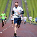Runner in a white t-shirt with number 702 competes in a run Stormont race on a red path, with grassy slopes and lamp posts lining the iconic course.