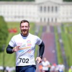 A man, donning a "The CCU Stormont Mile" T-shirt and bib number 702, races energetically past the grand Stormont building and its expansive lawn.