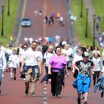 A group of people walking on a wide road during the Stormont run, with some participants wearing themed t-shirts. The background shows more walkers and greenery, capturing the vibrant spirit of the event.