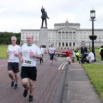 Participants are eagerly running in a charity race near the grand historic Stormont building, with a statue standing proudly in front.