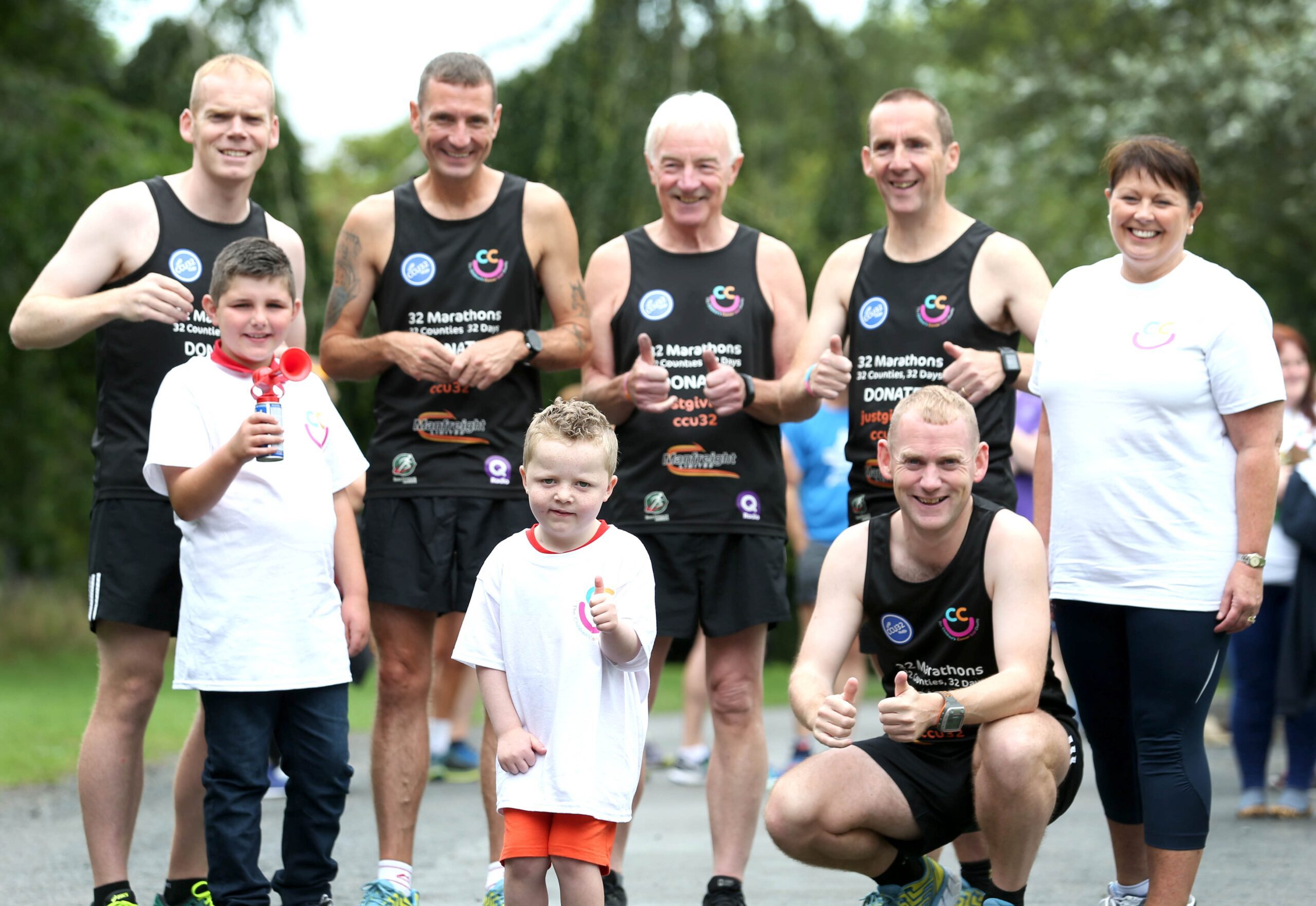 A group of Belfast runners in black athletic gear pose with two children and a woman in casual wear, giving thumbs up outdoors, celebrating their latest feat: the 32 County Challenge.