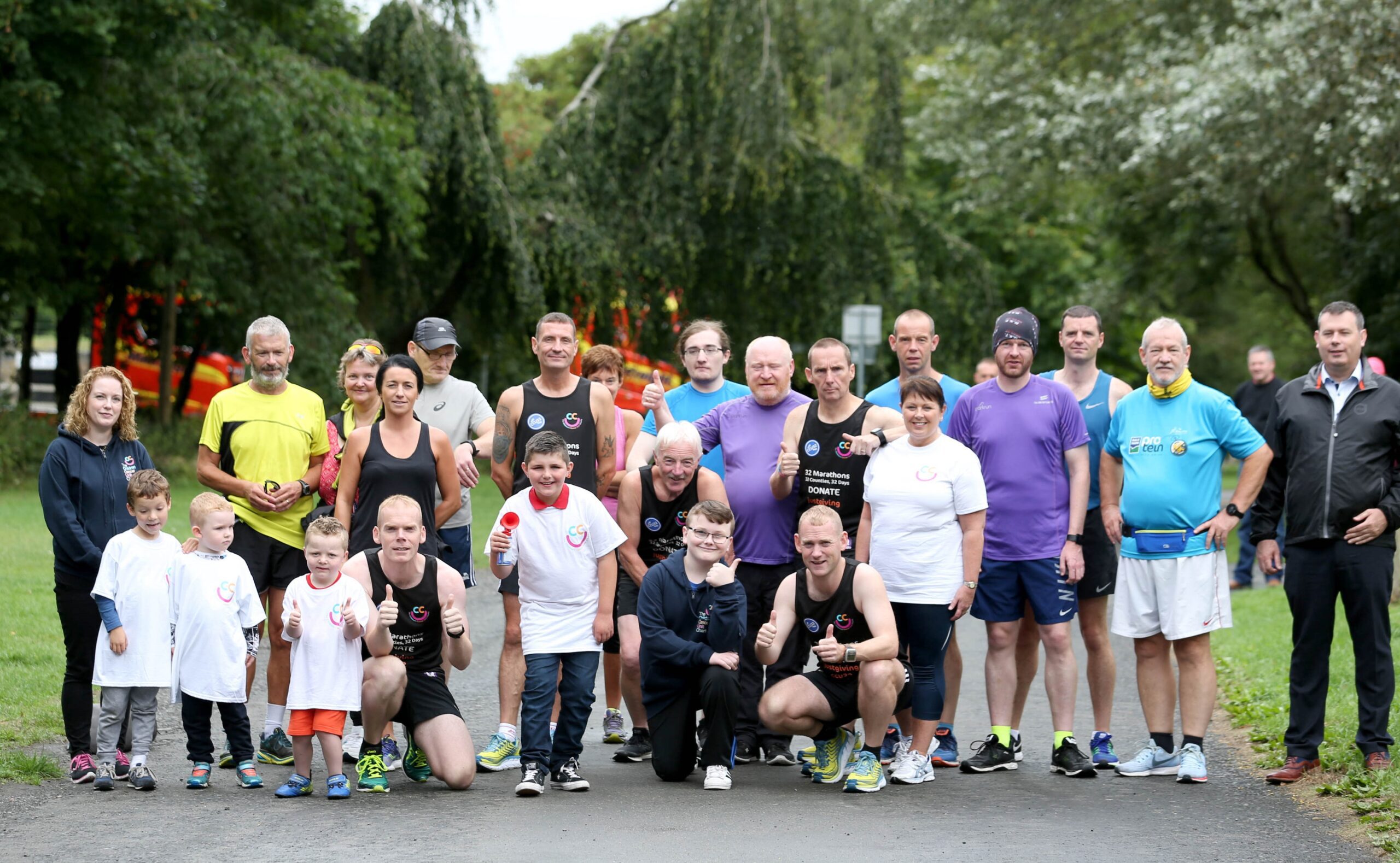 A group of Belfast runners, including adults and children, pose outdoors in athletic clothing. They appear ready for the 32 county challenge or event in a park setting with trees in the background.