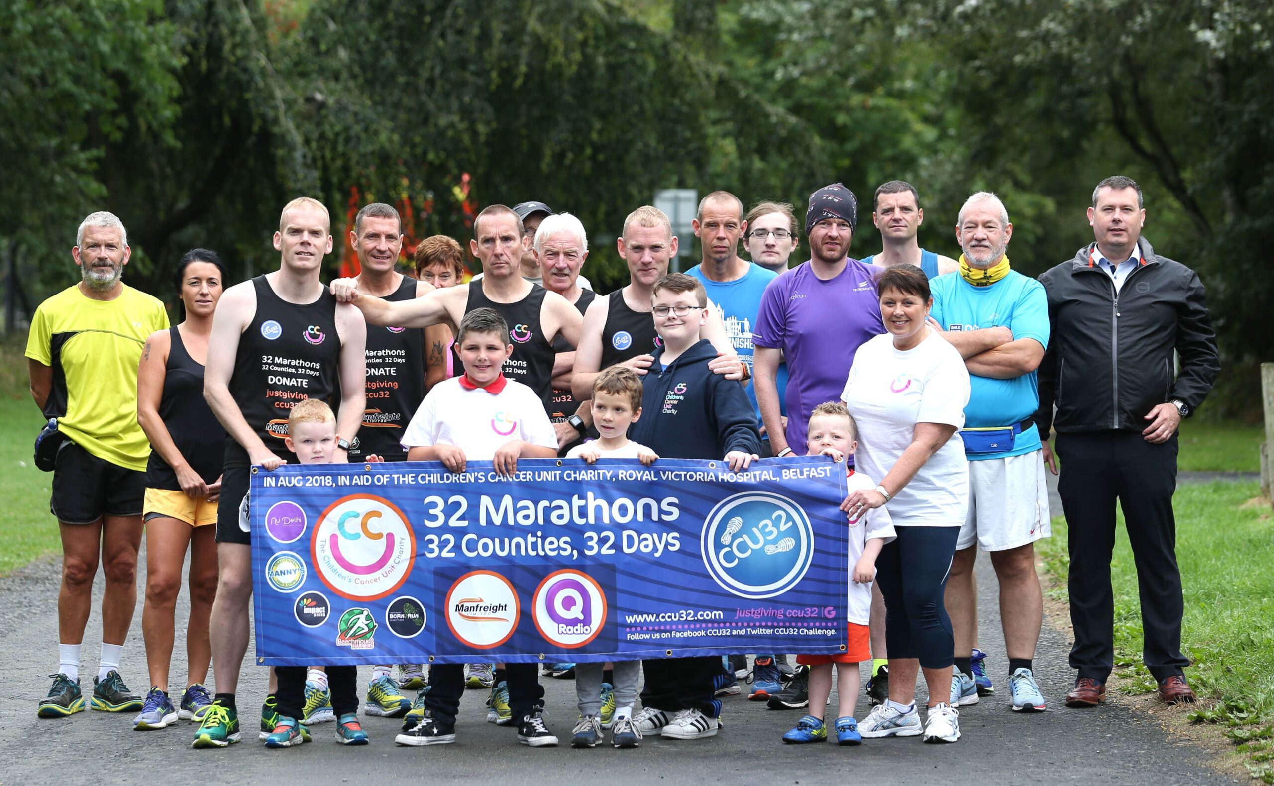 A group of Belfast runners proudly pose outdoors with a banner that reads "32 Marathons, 32 Counties, 32 Days," highlighting their incredible 32 county challenge and featuring various sponsor logos.
