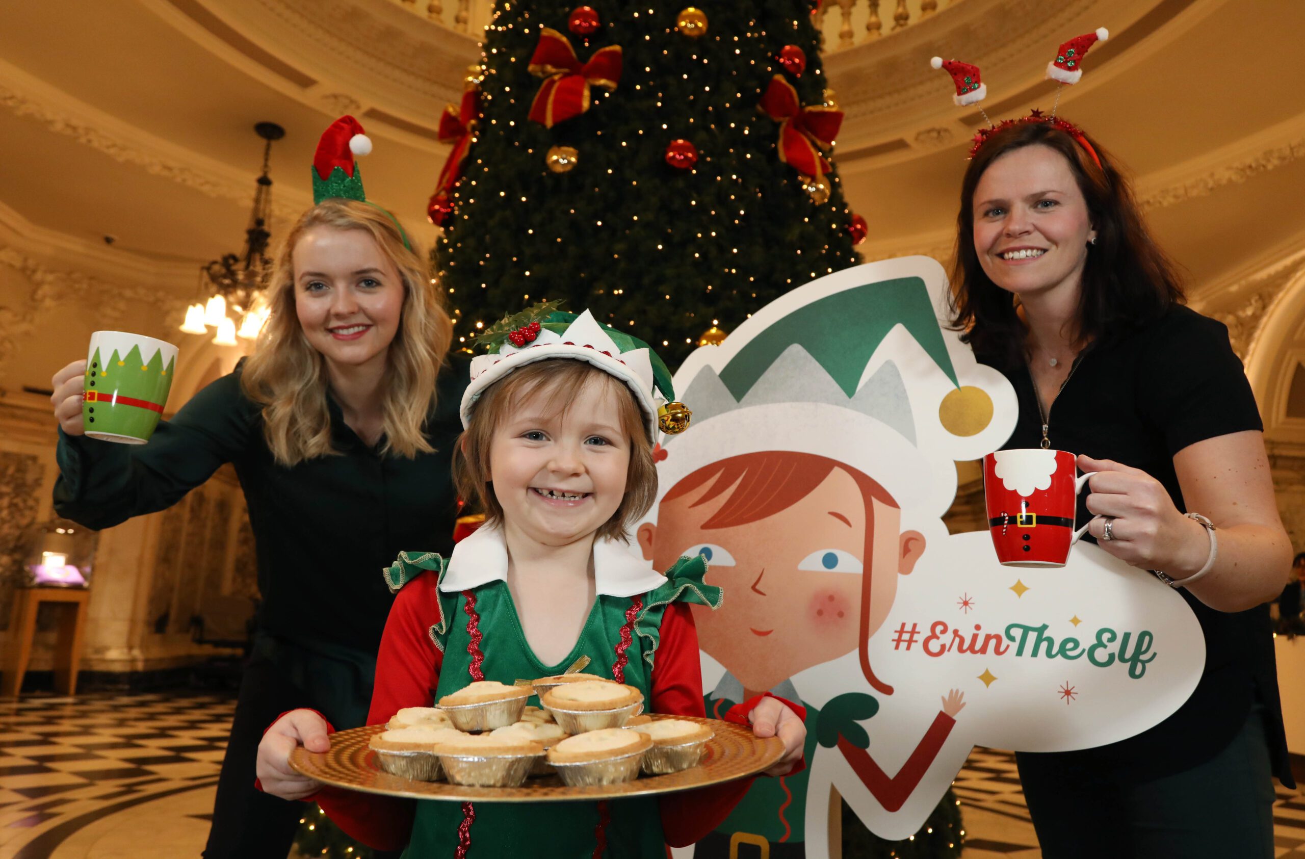 Three people in festive attire stand by a large Christmas tree. The child in front holds a tray of mince pies, while two adults hold mugs. A cardboard elf cutout is beside them.