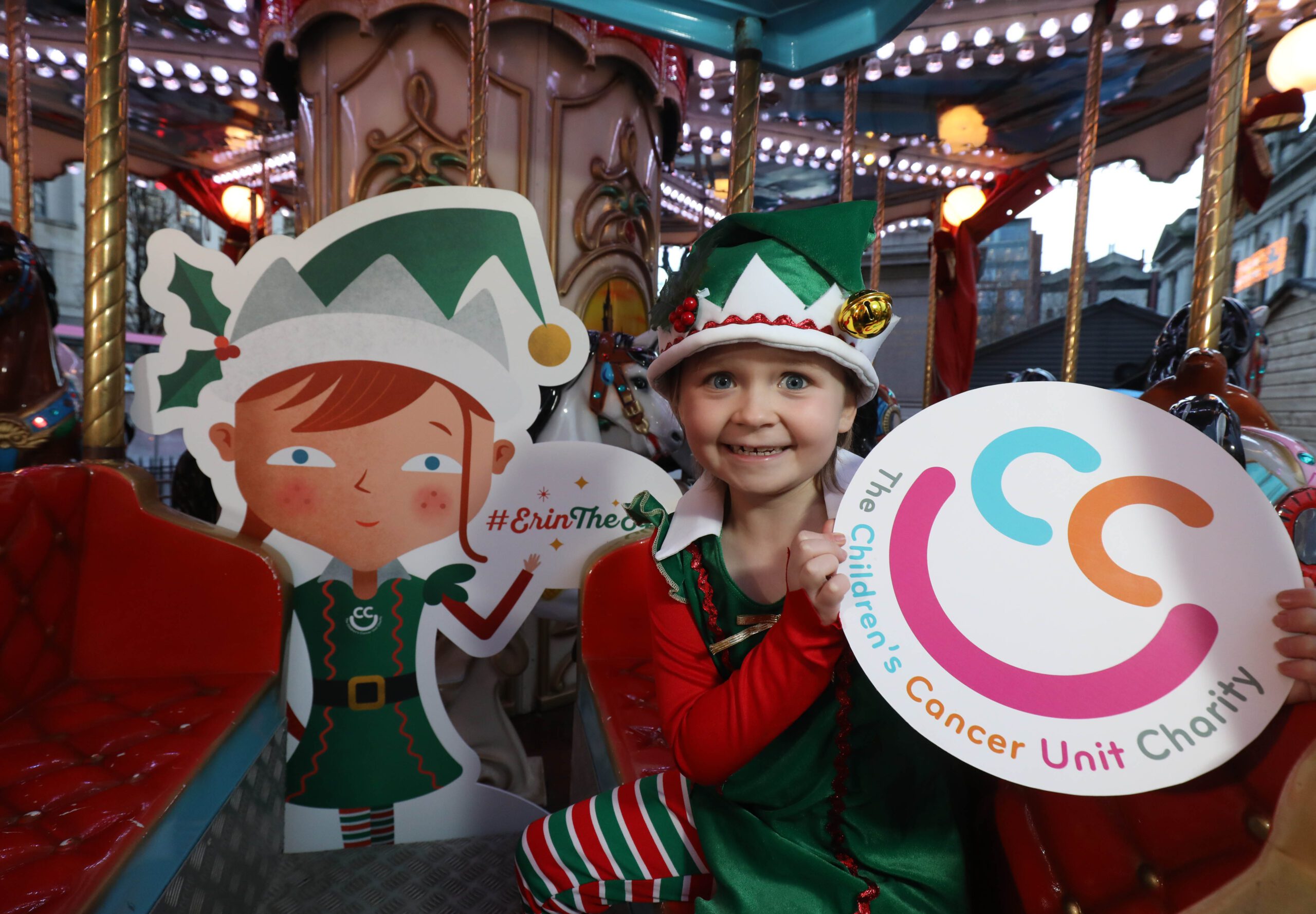 Child in elf costume holding a sign with the Children's Cancer Unit Charity logo, sitting near an elf cutout on a carousel.