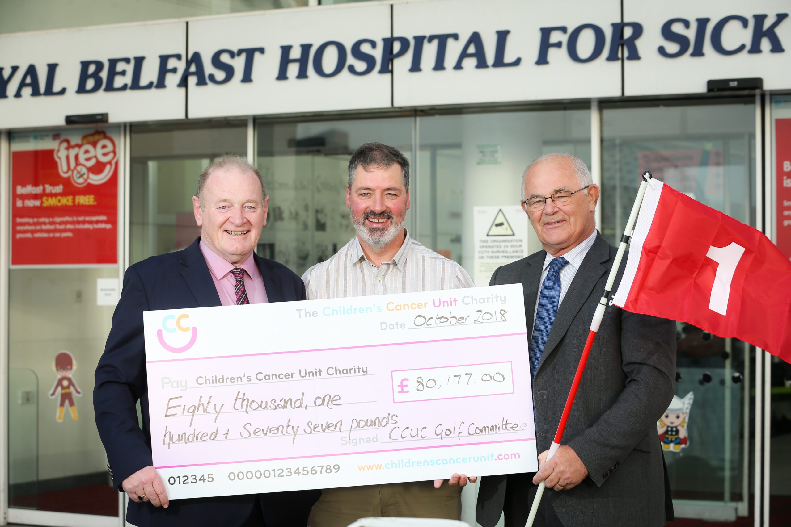 Outside the Royal Belfast Hospital for Sick Children, three men proudly present a large check for £80,177 to support the Children’s Cancer Unit. One of them holds a red flag, symbolizing the Business Community Club's commitment to this vital cause.