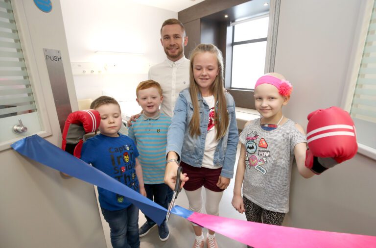 A group of cheerful children and an adult gather in a room. One child cuts a blue ribbon with scissors while others smile, and two kids wear boxing gloves, supporting their friends in the new children's cancer care facility.