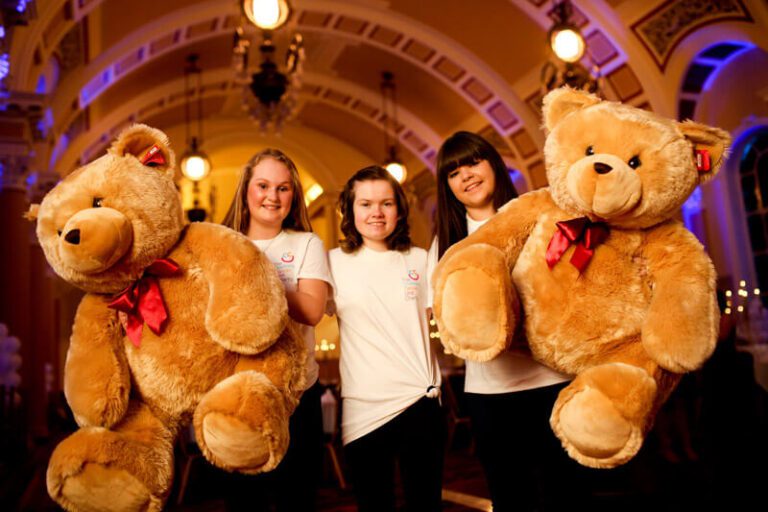 Three people in white shirts hold large teddy bears with red bows, standing in a decorated hall with arched ceilings and hanging lights, as part of the Teddies for Charity event dedicated to raising funds for a worthy cause.