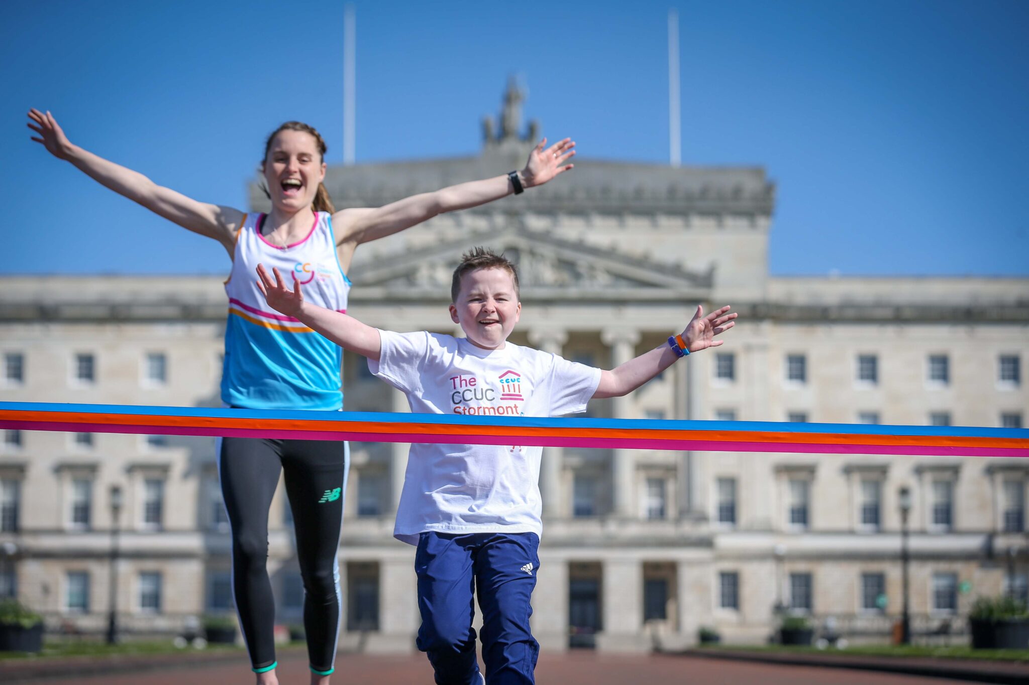 A young boy and a woman, inspired by Ciara Mageean, joyfully cross a red finish line in front of a large white building on a clear day.