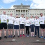 A group of ten people in matching event shirts stand in a line, posing for a photo in front of the grand Stormont building. They each wear numbered race bibs.
