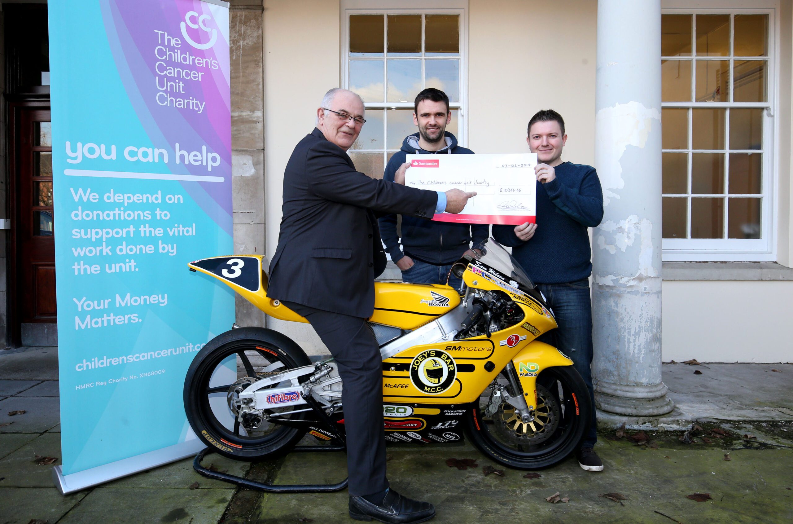 A group of four adults and one child proudly pose with a small yellow motorcycle in front of a building labeled "The Royal Belfast Hospital," symbolizing their shared goals to support the Children's Cancer Unit.