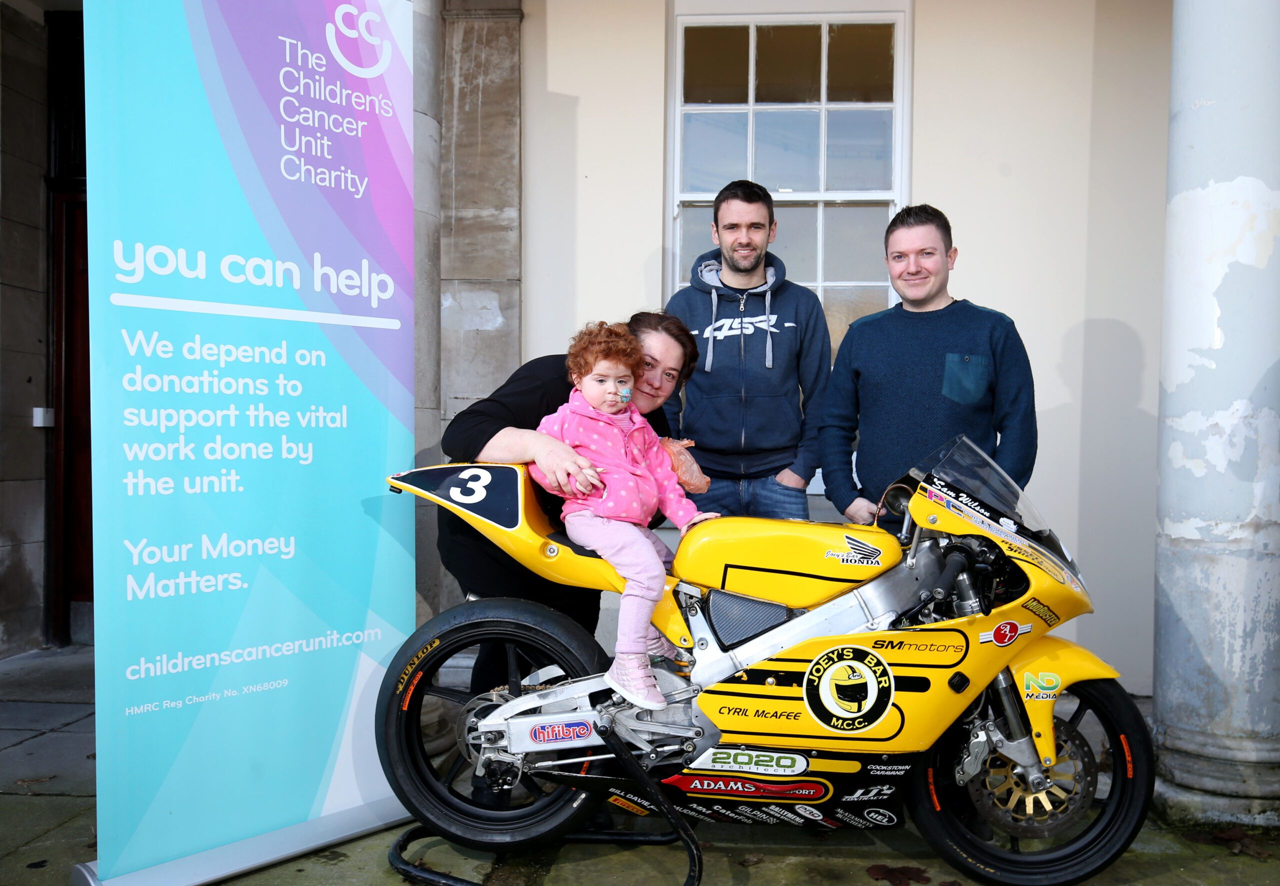A group of four adults and one child proudly pose with a small yellow motorcycle in front of a building labeled "The Royal Belfast Hospital," symbolizing their shared goals to support the Children's Cancer Unit.