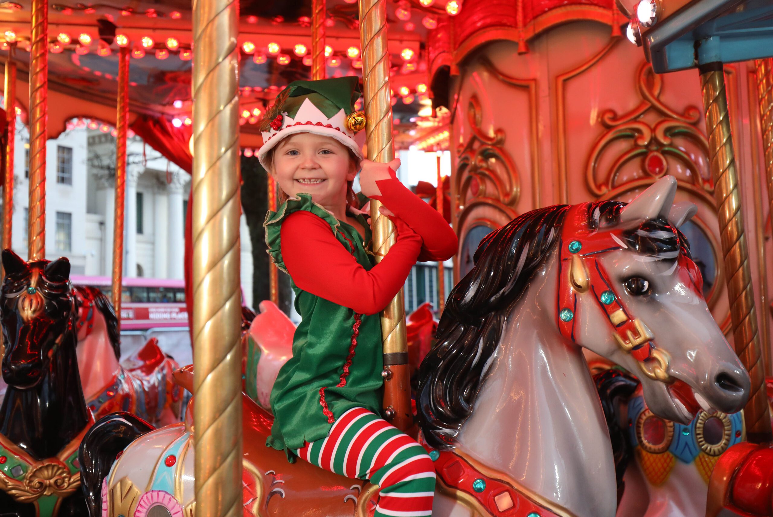 Smiling child dressed as Erin the Elf rides a carousel horse adorned with bright lights and festive decorations, capturing the joyful spirit of the Christmas Campaign.