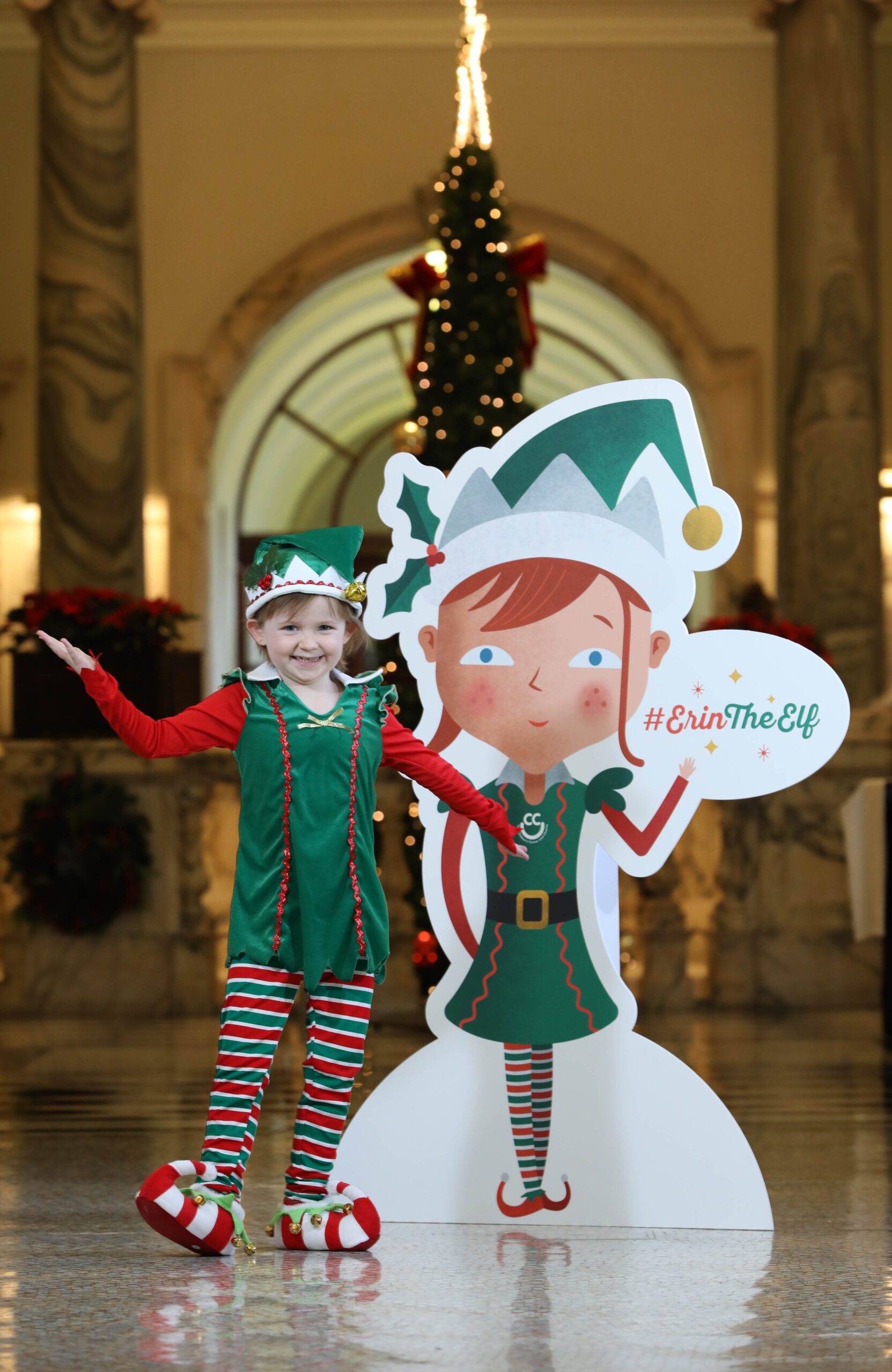 A child in a festive elf costume stands beside a cardboard cutout of an elf holding a sign that reads "#ErinTheElf." A decorated Christmas tree is visible in the background.