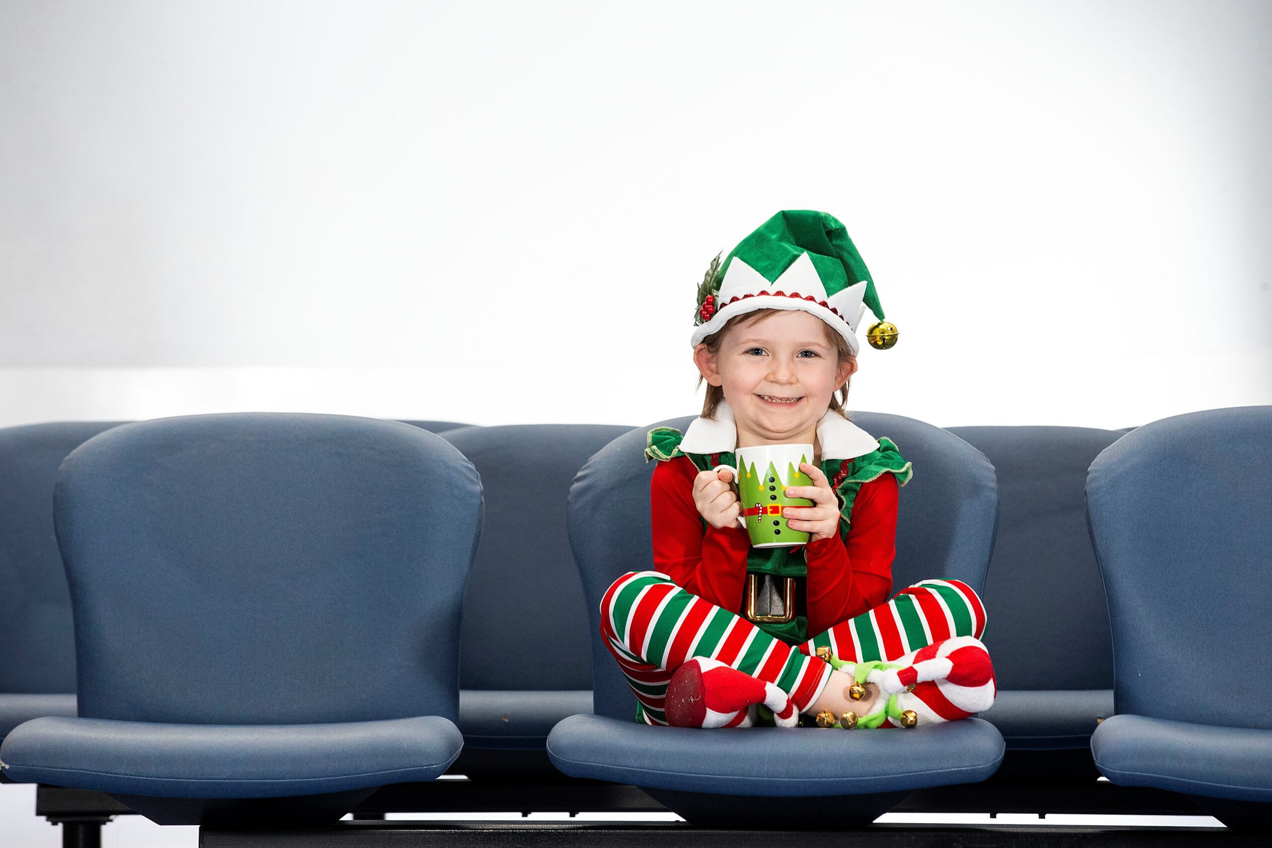 Child in elf costume sits cross-legged on chair, holding a mug, and smiling.