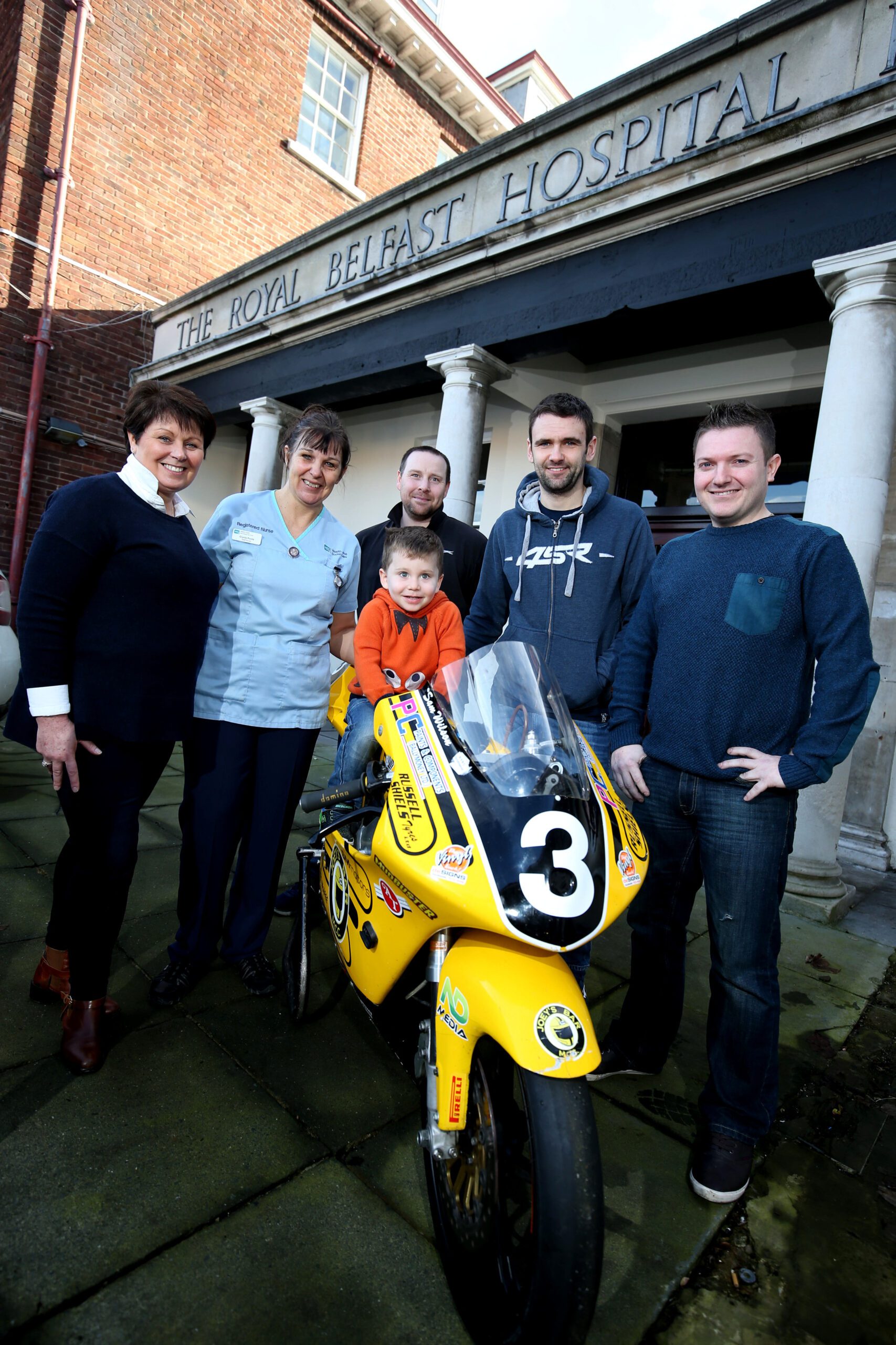 A group of four adults and one child proudly pose with a small yellow motorcycle in front of a building labeled "The Royal Belfast Hospital," symbolizing their shared goals to support the Children's Cancer Unit.