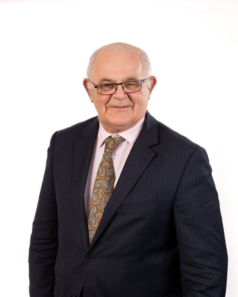 An esteemed member of our board, an elderly man in a dark pinstripe suit, light pink shirt, and paisley tie, smiles warmly at the camera against a plain white background.
