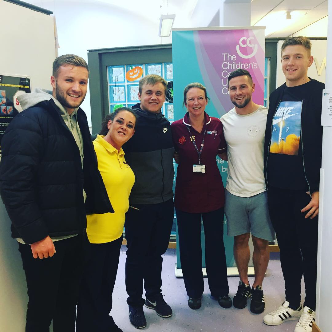 A group of six people stands together in a room, smiling warmly. Behind them, a banner for The Children's Cancer Charity is proudly displayed, hinting at their recent visit to the Children's Cancer Unit alongside representatives from Ulster Rugby.