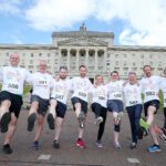 A group of people in matching event shirts, each with a number bib, pose with one leg raised triumphantly before the grand façade of Stormont.