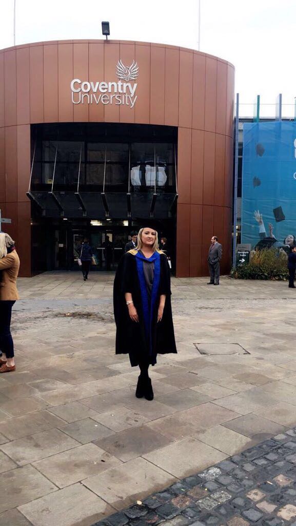 Michaela O'Kane, a proud graduate in cap and gown, stands beaming in front of the Coventry University building.